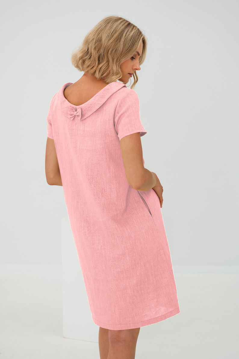 May include: A pink linen dress with a large collar and a bow detail on the back. The dress has short sleeves and a straight silhouette.