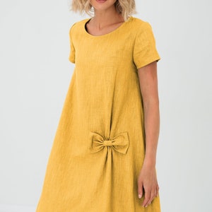 Könnte beinhalten: Senfgelbes Leinenkleid mit Schleifendetail. Das Kleid hat einen runden Ausschnitt und kurze Ärmel. Das knielange Kleid ist aus einem strukturierten Stoff gefertigt.
