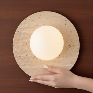 May include: A round, beige stone lamp with a glowing white orb in the center. The lamp is set against a dark brown background. The lamp is held by a hand with manicured nails.