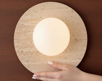 May include: A round, beige stone lamp with a glowing white orb in the center. The lamp is set against a dark brown background. The lamp is held by a hand with manicured nails.