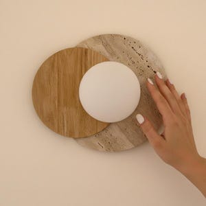 May include: A modern wall sconce featuring a white globe light, set against a backdrop of a light brown wooden circle and a beige stone circle. The design is minimalist and contemporary, suitable for home decor.