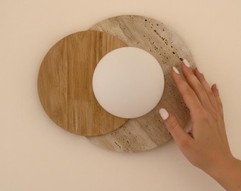May include: A modern wall sconce featuring a white globe light, set against a backdrop of a light brown wooden circle and a beige stone circle. The design is minimalist and contemporary, suitable for home decor.