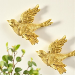 Op de afbeelding: Twee goudkleurige vogelmuursculpturen in vlucht. De vogels hebben gedetailleerde veertjesstructuren en zijn bevestigd op een lichtgekleurde muur. De sculpturen zijn gepositioneerd in de buurt van groen gebladerte, wat wijst op een door de natuur geïnspireerd thema.