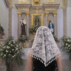 Pu&ograve; includere: Un velo di pizzo bianco con un motivo floreale e un bordo smerlato, indossato da una persona in chiesa. Lo sfondo presenta statue religiose, dipinti e composizioni floreali, con dettagli dorati.