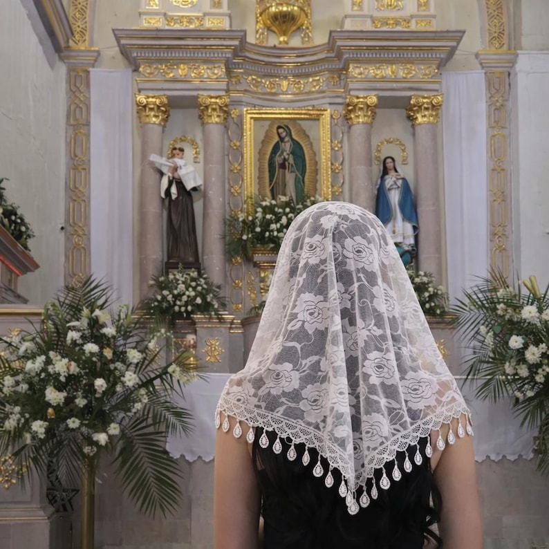 Pu&ograve; includere: Un velo di pizzo bianco con un motivo floreale e un bordo smerlato, indossato da una persona in chiesa. Lo sfondo presenta statue religiose, dipinti e composizioni floreali, con dettagli dorati.
