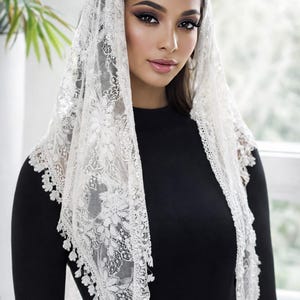 May include: A white lace veil featuring floral designs and a scalloped trim. The veil is worn over a black top, creating a contrast of textures and colours. The delicate lace adds a touch of elegance.