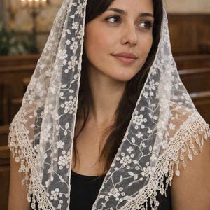 Può includere: Un velo di pizzo bianco con ricami floreali e bordo smerlato. Il velo è drappeggiato sulla testa e sulle spalle, con una delicata frangia sul fondo. Lo sfondo è un interno di chiesa.