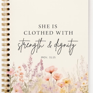 May include: Cream-coloured spiral-bound notebook. The cover reads "SHE IS CLOTHED WITH strength & dignity" and "PROV. 31:25." A watercolour floral design in pink, peach, and purple adorns the bottom of the cover. The notebook has a gold spiral binding.