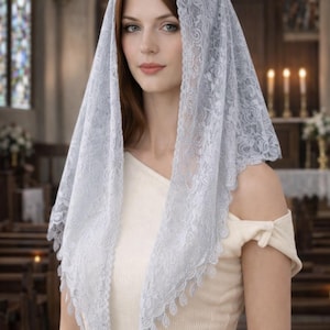 Pu&ograve; includere: Un velo di pizzo azzurro con un motivo floreale. Il velo &egrave; drappeggiato sulla testa e sulle spalle. La persona indossa un top color crema. Lo sfondo &egrave; un interno di chiesa con vetrate.