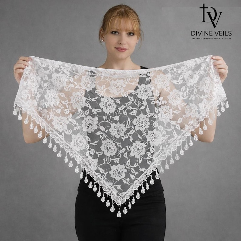 Pu&ograve; includere: Una mantilla di pizzo bianco con un motivo floreale a rose e un bordo smerlato con dettagli a goccia. Il logo Divine Veils &egrave; visibile in alto a destra. La mantilla &egrave; tenuta da una persona.