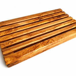 May include: A rectangular wooden trivet constructed from six parallel, light brown olive wood slats with a distinct natural grain pattern. The slats are joined together to form a flat, slatted surface with narrow gaps between each individual piece of wood.
