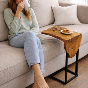 May include: A live edge side table with a natural wood top and black metal base. The table is next to a sofa. A plate of cookies and a pair of glasses sit on the table. The table is approximately 24 inches long.