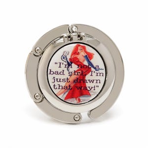 May include: Silver-toned purse hook with a circular design. The center features a graphic of a cartoon woman in a red dress and the quote, "I'm not a bad girl, I'm just drawn that way!"