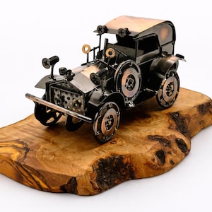 May include: A handcrafted metal model of a vintage car, featuring a black body, silver accents, and copper-colored wheels. The car sits on a natural wood base, showcasing the intricate details of the miniature vehicle.