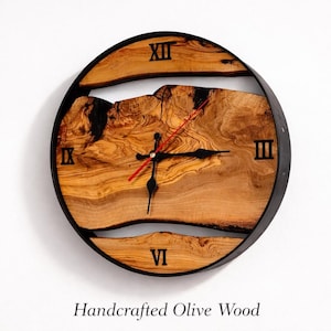 May include: A handcrafted olive wood wall clock with black Roman numerals and hands. The clock has a natural wood grain design and a black metal frame. The second hand is red. The text "Handcrafted Olive Wood" is at the bottom.