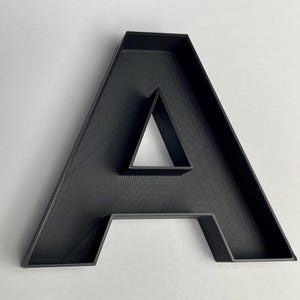 Pode incluir: Uma letra A tridimensional com acabamento em preto fosco. A letra apresenta uma estrutura oca tipo bandeja com bordas elevadas e uma abertura triangular distinta no centro, exibindo uma superfície texturizada típica de objetos impressos em 3D.