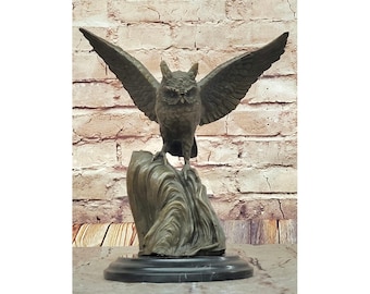 May include: A bronze owl sculpture with outstretched wings, perched atop a textured rock formation. The owl's detailed feathers and facial features are visible. The sculpture sits on a round, black base, against a brick wall background.