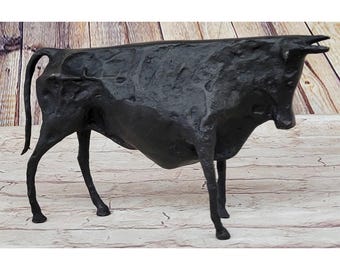 Puede incluir: Una escultura de bronce oscuro de un toro en posición de pie. El toro tiene una superficie texturizada y está posicionado con la cabeza ligeramente baja. La escultura está sobre un fondo de tablones de madera.