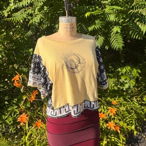 May include: A mustard yellow short-sleeve shirt featuring a black line-art graphic of a sun with a face on the chest. The sleeves and the bottom hem are accented with a black and white patterned fabric featuring geometric and floral motifs.