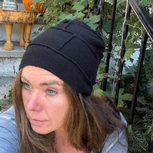 May include: A black beanie hat featuring a paneled construction with visible stitched seams. The hat has a ribbed knit band at the base and a soft, solid black fabric crown that fits snugly around the head, providing full coverage.