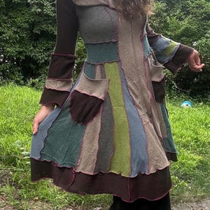 May include: A hooded coat featuring a patchwork design with various fabric panels in shades of brown, green, blue, and gray. The garment includes a front zipper closure, long sleeves with flared cuffs, and two large patch pockets on the front.