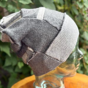 May include: A slouchy beanie hat constructed from multiple panels of knit fabric in varying shades of gray, including charcoal, medium gray, and light gray, featuring visible stitched seams and a wide ribbed knit band at the base.