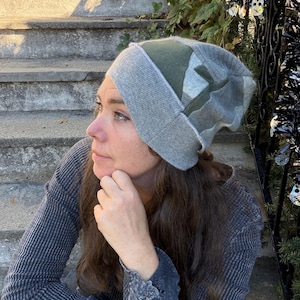 May include: A slouchy beanie hat constructed from panels of fabric in shades of heather gray and olive green. The hat features a wide, ribbed knit brim and a soft, unstructured crown with visible seams connecting the different colored fabric sections.