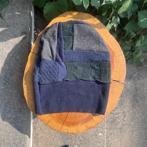 May include: A beanie hat constructed from various fabric patches in shades of navy blue, charcoal gray, and dark forest green. The design features a mix of smooth knit and cable knit textures, finished with a wide, ribbed navy blue hem at the base.