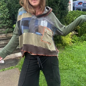 Puede incluir: Una sudadera con capucha y manga larga que presenta un diseño de patchwork con paneles de tela en color verde oliva, azul y gris. La prenda incluye un bolsillo en el pecho con cierre de botón, un dobladillo acanalado marrón y costuras naranjas.