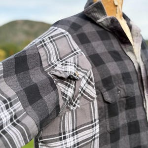 May include: A long-sleeve button-up shirt featuring a patchwork design of grey and black plaid patterns. The garment includes a collar, a front chest pocket, and long sleeves, displayed on a mannequin torso against an outdoor background.