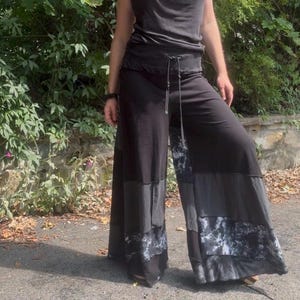 May include: Black wide-leg pants featuring a drawstring waist and a patchwork design. The lower sections of the pant legs incorporate panels of solid black fabric alternating with dark gray and black tie-dye patterned fabric with raw edges.