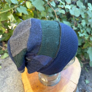 May include: A slouchy beanie hat constructed from multiple panels of knit fabric in shades of charcoal gray, navy blue, and dark forest green. The design features a mix of smooth jersey knit and textured cable knit patterns throughout the piece.