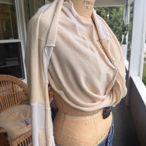 May include: A cream-colored long-sleeve knit garment featuring a draped cowl neckline and contrasting white panels along the sleeves and collar. The piece is displayed on a beige dress form, showcasing a soft, lightweight texture and a loose fit.