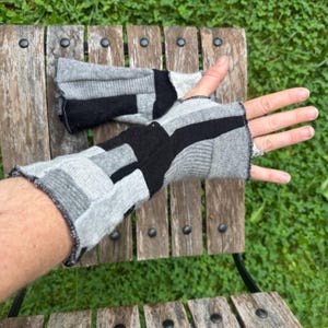 Könnte beinhalten: Ein Paar fingerlose Handschuhe mit einem Patchwork-Design aus grauem und schwarzem Strickstoff. Die Handschuhe haben eine gerippte Textur und rohe, offene Kanten, wobei ein Handschuh an der Hand getragen wird und der andere flach auf Holz liegt.