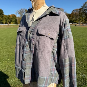 May include: A long-sleeve button-up shirt featuring a patchwork design of solid grey fabric and a green, blue, and orange plaid pattern. The shirt includes a classic collar, two chest pockets with buttoned flaps, and a front button closure.