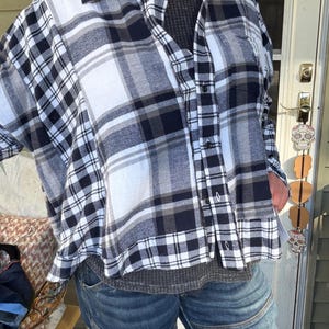 May include: A long-sleeve button-down shirt featuring a black, white, and gray plaid pattern. The shirt has a collar, a front button placket, and a single chest pocket. It is worn over a dark gray textured shirt and blue denim jeans.