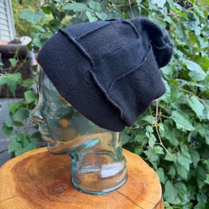 May include: A black beanie hat featuring a structured, panelled design with visible seams. The knit fabric has a soft texture and a fitted silhouette, designed to sit snugly on the head with a slightly slouchy crown at the back.
