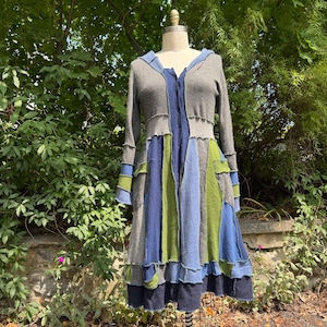 May include: A long-sleeved, knee-length cardigan featuring a patchwork design of grey, blue, and green knit fabric panels. The garment has a hood, a button-front closure, and a flared silhouette with visible seams connecting the various colored sections.