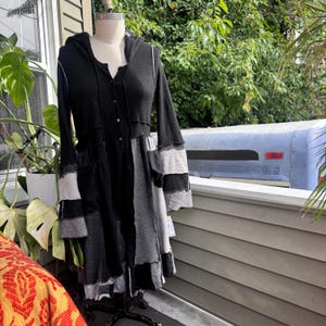 Puede incluir: Un vestido con capucha y manga larga que presenta un frente abotonado y un diseño de retazos. La prenda está confeccionada con paneles de tela en negro, gris carbón y gris claro con costuras visibles de borde sin rematar en toda la pieza.