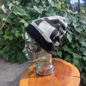 May include: A knit beanie featuring a checkered pattern in shades of black and light gray. The hat has a solid black ribbed cuff at the base and a slouchy, relaxed fit. The fabric displays a soft, woven texture throughout the crown and body.