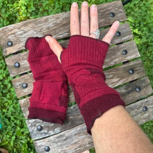 May include: A pair of deep burgundy fingerless gloves featuring a ribbed knit texture. The gloves extend from the knuckles down to the wrist, showcasing a consistent dark red color throughout the fabric with a slightly darker ribbed cuff edge.