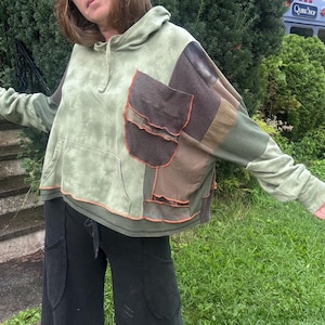 Könnte beinhalten: Ein langärmeliger Kapuzenpullover mit einer salbeigrünen Batik-Basis und kontrastierenden braunen und dunkelbraunen Stoffbahnen an der Seite und am Ärmel. Das Kleidungsstück verfügt über eine große Vordertasche und sichtbare orangefarbene Nähte.