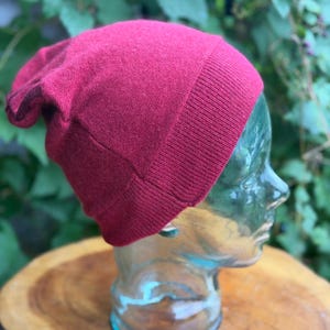 May include: A deep red beanie hat with a fine knit texture and a wide, ribbed cuff at the base. The hat features a slightly slouchy fit and is displayed on a clear, transparent mannequin head.