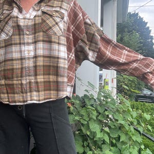 May include: A long-sleeve button-down shirt featuring a patchwork design of brown, tan, and white plaid fabric. The shirt includes two chest pockets with flaps, a standard collar, and light-colored buttons running down the center front placket.