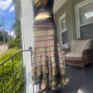 May include: A long, floor-length skirt featuring a patchwork design of horizontal fabric strips in various shades of olive green, brown, and tan. The skirt has a wide, solid dark brown waistband and visible seams between each horizontal fabric panel.
