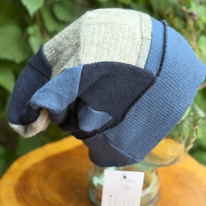 May include: A slouchy beanie hat constructed from a patchwork of knit fabric panels in shades of heather gray, navy blue, and medium blue. The hat features a wide, ribbed blue cuff at the base and includes a small white rectangular product tag.