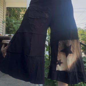 Op de afbeelding: Zwarte broek met wijde pijpen, hoge taille, een grote opgestikte zak op de rechterheup en een gelaagde constructie. Het linker onderbeen heeft een gebleekt rechthoekig paneel met twee donkere sterren en een afwerking met een ruchezoom.
