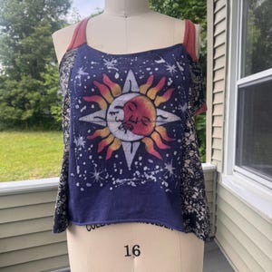 May include: A sleeveless tank top featuring a central graphic of a sun and moon face against a dark blue background with white star accents. The garment has solid rust-colored straps and side panels with a black and white floral pattern.