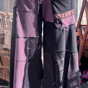 May include: Wide-leg pants featuring a patchwork design in black and muted purple. The garment includes waffle-knit fabric panels, a front pocket, and white line-art illustrations of faces and geometric patterns on the lower leg sections.