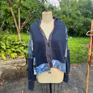 May include: A navy blue zip-up hoodie featuring a hood, long sleeves, and a front kangaroo pocket. The garment is accented with exposed orange stitching along the seams and a blue and white tie-dye pattern on the lower hem and sleeve panels.